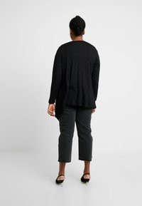 Kobieta z warkoczami, ubrana w czarną bluzkę z długim rękawem, ciemne jeansy 3/4 i czarne szpilki, stojąca na tle białym.