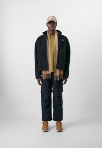 Schwarze Fleecejacke, olivgrünes Hemd, brauner Schal mit Fransen, schwarze Cargo-Hosen und braune Sneakers, topped mit einer beigen Kappe.