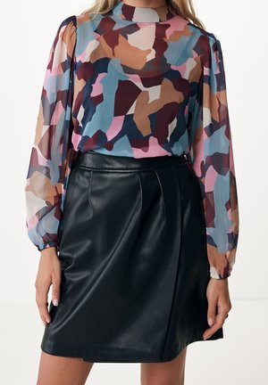 Femme portant un chemisier transparent à imprimé abstrait multicolore et une jupe en cuir noire, debout, les mains détendues le long du corps.