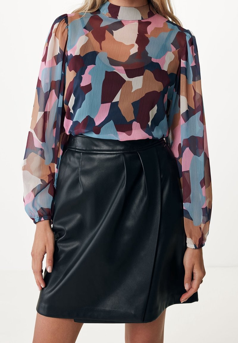 Femme portant un chemisier transparent à imprimé abstrait multicolore et une jupe en cuir noire, debout, les mains détendues le long du corps.