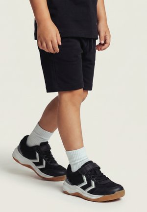 Barn iført sorte shorts, sort/hvide sneakers med hvide sokker, stående med det ene ben let fremad på en lys baggrund.