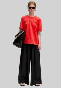 Rotes Baumwoll-T-Shirt, schwarze weite Hosen, übergroße schwarze Tasche und Sonnenbrille mit eingeschränkter Sicht. Glatte Texturen und kräftige Farben hervorgehoben.