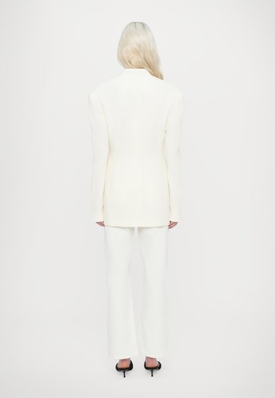 Witte op maat gemaakte blazer met brede schouders, gecombineerd met bijpassende witte uitlopende broeken en zwarte heeled sandalen. Soepele stofstructuur.