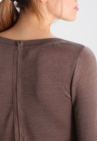 Brauner Pullover mit glatter Textur, der über einen horizontalen Ausschnitt und einen dezenten Reißverschluss am Rücken verfügt. Einfaches und körpernahes Design.