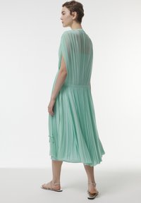 Vestido plisado color verde menta con mangas cortas, cintura ceñida y falda fluida. Presenta lazos en los laterales y una textura ligera y transparente.