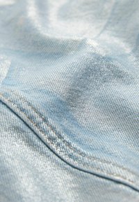 Tissu en denim bleu clair avec une surface brillante et texturée, présentant des coutures visibles et une apparence lisse légèrement froissée.