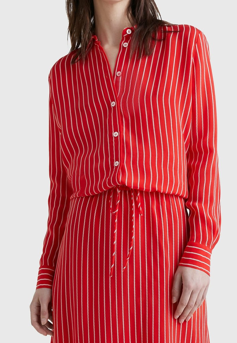 Femme portant une robe rouge à manches longues avec des rayures verticales blanches, boutonnée devant et une taille cintrée par un cordon.