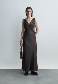 Žena v tmavohnedých saténových šatách bez rukávov s čipkovaným lemom nesie malú bordovú kabelku, stojaca pred jednoduchou sivou stenou.