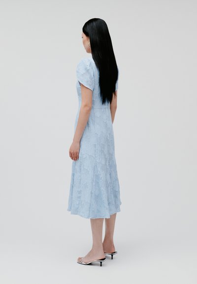 Hellblaues Midi-Kleid mit strukturiertem Blumendessin, kurzärmlig und mit ausgestelltem Saum; kombiniert mit silbernen Absatzsandalen.