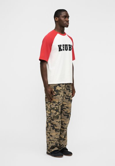 Λευκό μπλουζάκι με κόκκινα μανίκια ρ άγκλαν, με μαύρο κείμενο "KSUBI". Συνδυασμένο με μπεζ παραλλαγής παντελόνια cargo και μαύρα sneakers.