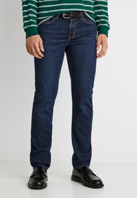 Άνδρας που φοράει στενά σκουρό μπλε τζιν, μαύρα δερμάτινα παπούτσια και πράσινο-λευκό ριγέ πουκάμισο με μακριά μανίκια, όρθιος μπροστά σε ανοιχτό φόντο.