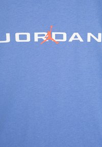 Světle modré tričko s bílým nápisem "JORDAN" a oranžovým logem Jumpman ve středu. Vyrobeno z měkké bavlněné tkaniny.