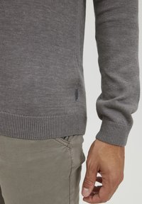 Grauer Pullover mit geripptem Saum und Bündchen, aus einem glatten Strickstoff gefertigt. Die Seitennaht zeigt ein kleines Etikett. Kombiniert mit hellgrauen Hosen.