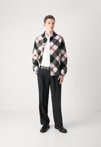 Plaid jacket in black, white, and red with a button closure, worn over a white shirt, paired with black trousers and shoes. Simple background.