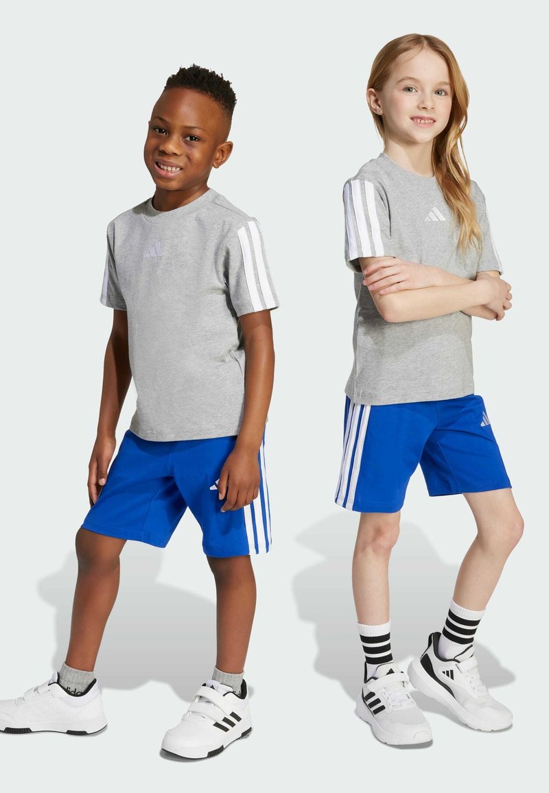 Camisa gris de manga corta con rayas blancas y logo, combinada con pantalones cortos azules con rayas blancas. Ambos llevan zapatillas deportivas blancas.