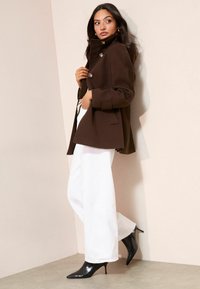 Rudas vilnos mišinys paltas su aukštu apykakliu, auksiniais mygtukais, derinamas su baltomis platėjantėmis kelnėmis ir juodais aukštakulniais su aštriu nosimi, neutraliame fone.