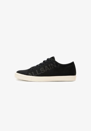 Zapatillas de lona negras con suela de goma color crema, que presentan ojales y cordones negros, y un logo sutil impreso en el costado.