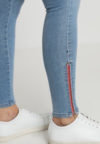 Jeans skinny de color azul claro con cremalleras rojas en el dobladillo del tobillo, hechos de tela de mezclilla; combinados con zapatillas blancas sobre un fondo neutro.