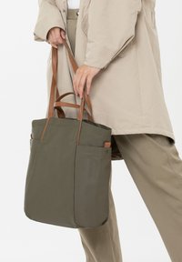 Persoon in een beige outfit die een olijfgroene totebag met bruin leren handvatten vasthoudt, staand tegen een witte achtergrond.
