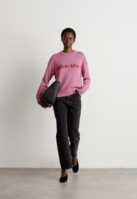 Maglione rosa con logo "calvin klein", pantaloni neri, tiene una borsa a mano nera, scarpe nere, su uno sfondo neutro.