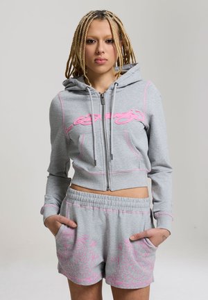 Młoda osoba z blond warkoczami nosząca szarą krótka bluzę z kapturem z różowymi literami oraz dopasowane szare szorty z różowymi kwiatowymi wzorami.
