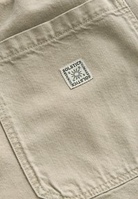 Beige kangas, jossa on hienovarainen, teksturoitu kudonta, jonka etiketissä on neliömuotoinen label, jossa lukee "SOLSTICE" ja aurinkokuvasto.