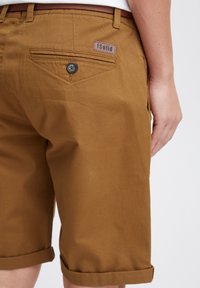 Brown cotton shorts with rolled cuffs and a buttoned back pocket. Features a branded leather patch. Smooth texture and tailored design.