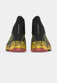 O pereche de pantofi sport negri și galbeni cu pete, cu talon roșu, văzuți din spate, având textul "URAGANO GTX" pe călcâie.