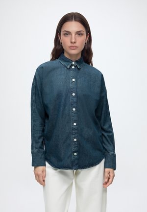 Kvinna med långt brunt hår som bär en mörkblå knäppt jeansskjorta och vita byxor, stående mot en enfärgad ljus bakgrund.