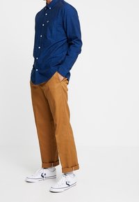 Mörkblå skjorta med knappar och bröstficka, i kombination med ljusbruna vida byxor och vita sneakers med blå detaljer.