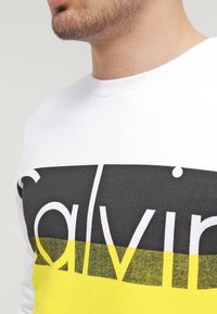 Sudadera blanca con un gran logo "calvin" en negro y amarillo. El escote es acanalado, con una textura suave y una forma ajustada.