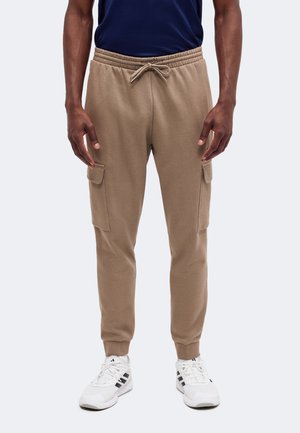 Άνδρας με μπεζ παντελόνι jogger cargo με κορδόνι στη μέση και λευκά αθλητικά παπούτσια σε μονόχρωμο φόντο.