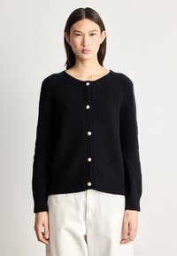 Juodas, pūkuotas kardiganas su apvalia iškirpte ir devyniais perlų pavidalo mygtukais per visą priekinę dalį. Ilgos rankovės ir laisvas siluetas.