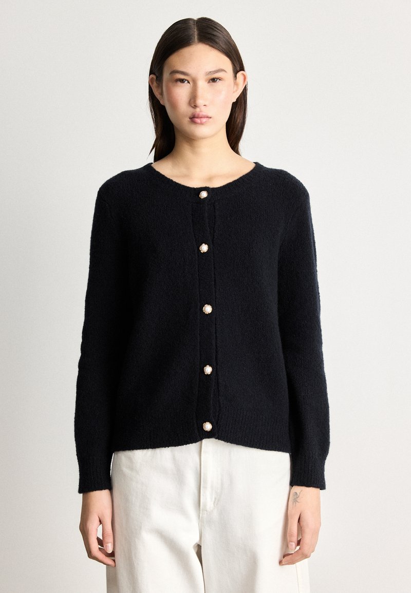 Juodas, pūkuotas kardiganas su apvalia iškirpte ir devyniais perlų pavidalo mygtukais per visą priekinę dalį. Ilgos rankovės ir laisvas siluetas.