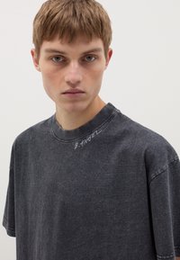 Camiseta de algodón de color gris oscuro, de corte oversized, con mangas cortas, cuello redondo y texto bordado "B.ANGEL" en el hombro izquierdo.