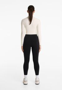 Fekete leggings magas derekú, egy testhez simuló, hosszú ujjú krémszínű toppal párosítva. Az öltözetet világosszürke sportcipő és fehér zokni egészíti ki.