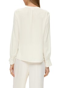 Blusa a maniche lunghe di colore crema, realizzata in tessuto leggero, con scollo rotondo, polsini con bottoni e orlo leggermente curvato.
