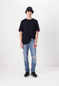 Marineblå t-shirt med rød og hvid kant, båret sammen med lyseblå slidte jeans og sorte sneakers. Modellen bærer en sort bækhue.