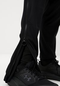 Calças atléticas pretas com fechos de correr laterais, feitas de um tecido suave. Combinadas com ténis pretos com pormenores texturizados e logótipo.