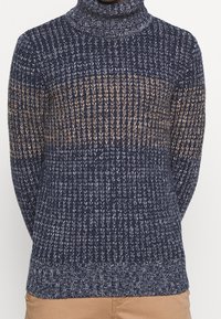 Pull en maille à col roulé en bleu marine avec des rayures beiges, présentant un motif texturé, des poignets côtelés et une coupe légèrement cintrée à la taille.