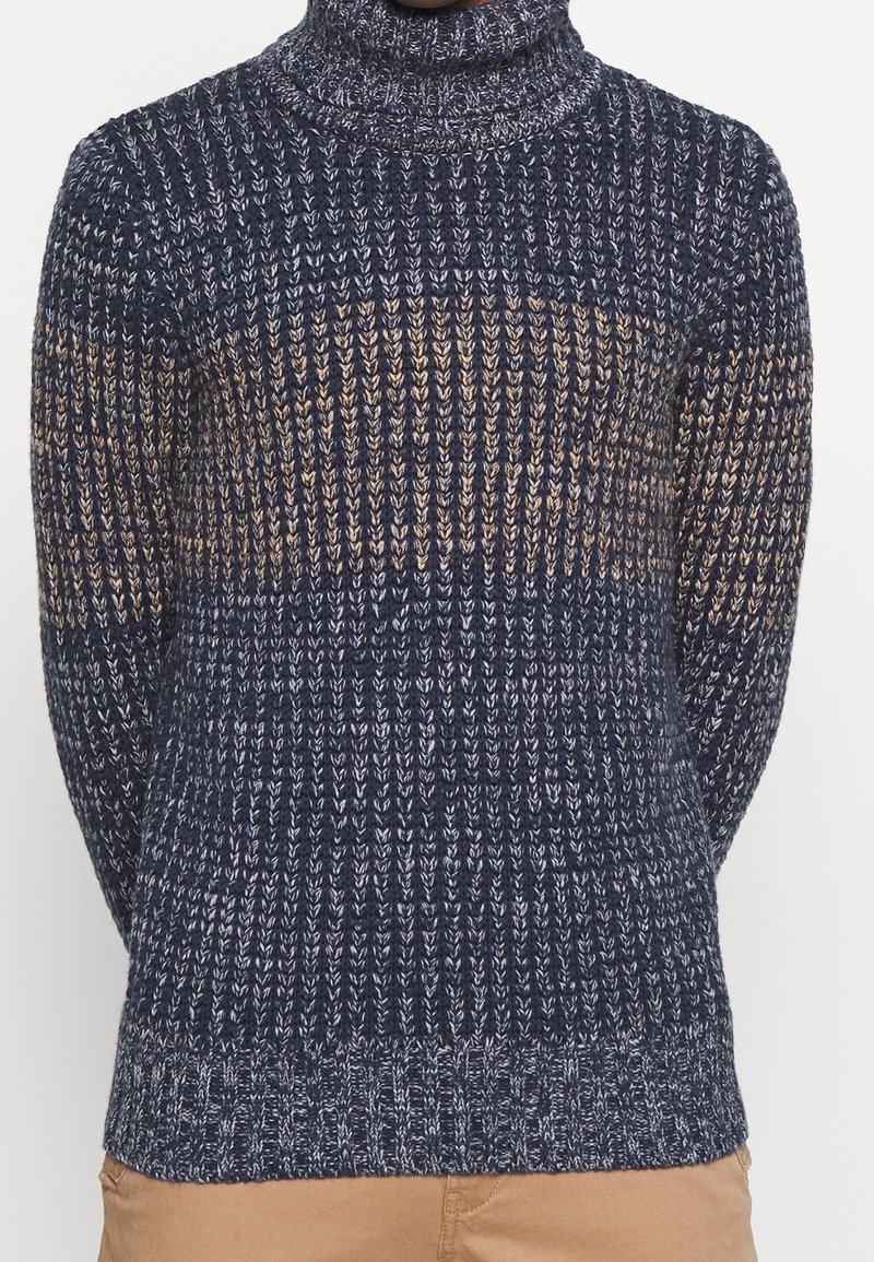 Pull en maille à col roulé en bleu marine avec des rayures beiges, présentant un motif texturé, des poignets côtelés et une coupe légèrement cintrée à la taille.