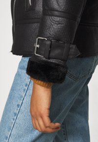 Manche de veste en cuir texturé noir avec sangle à boucle et poignet en fourrure sur une manche en tricot marron, associée à un jean en denim bleu clair.