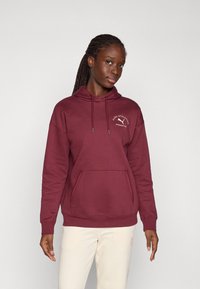 Burgunderfarget hettegenser laget av myk stoff, med en frontlomme, snorer og en liten logo detalj på brystet.