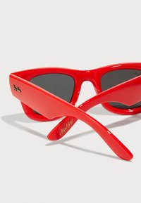 Røde plast solbriller med katteøyne-form, med mørke linser og "Ray-Ban"-logoen. Stengene har innskrift "AWGE" i gull.