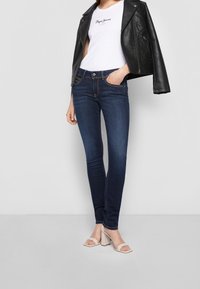 Mörkblå skinny jeans med medelhög midja, kombinerat med en vit åtsittande T-shirt och en svart lädermotorcykeljacka. Vita klacksandaler.