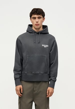 Junger Mann trägt einen dunkelgrauen Hoodie mit Fronttasche und „Service Works“-Logo auf der Brust, kombiniert mit olivgrünen Cargohosen.