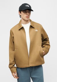 Νεαρός άνδρας φοράει μπεζ μπουφάν North Face, λευκό πουκάμισο, μπλε παντελόνι και μαύρο καπέλο, κοιτάζει προς τα πλάγια με το ένα χέρι στην τσέπη του μπουφάν.