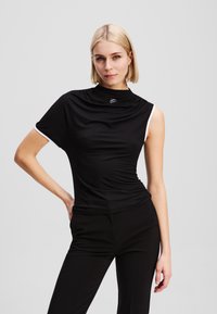 Melnas īsām piedurknēm tops ar plūstošu dekoltē, ar baltām akcentēm vienā plecā un piegulošu siluetu. Gluda auduma apdare.