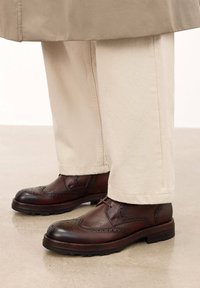 Zapatos de cuero marrón con detalles perforados, combinados con pantalones beige claros, de pie sobre una superficie neutra.