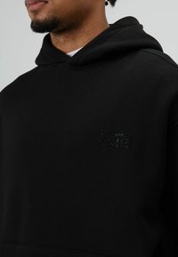 Schwarzer Kapuzenpullover aus weichem Stoff. Verfügt über ein strukturiertes Logo auf der Brust und eine geräumige Fronttasche.